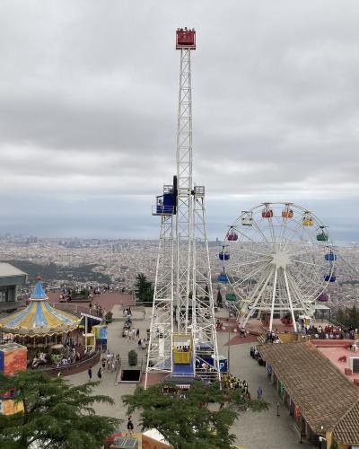An amusement park featuring a tall white drop tower ride, a large Ferris wheel, and a carousel, set on a hilltop overlooking a dense urban cityscape and the sea.