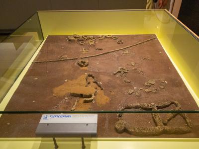 A museum display case containing a sand and stone diorama of ancient structures, with a label reading "FUERTEVENTURA" and a logo for "MUSEO DE LA NATURALEZA Y EL HOMBRE".