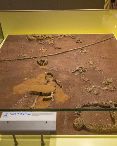 A museum display case containing a sand and stone diorama of ancient structures, with a label reading "FUERTEVENTURA" and a logo for "MUSEO DE LA NATURALEZA Y EL HOMBRE".
