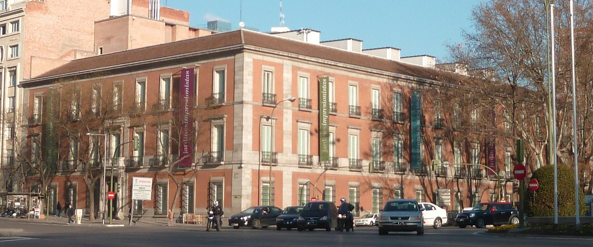 A street-level view of a large, multi-story brick building with vertical banners hanging from the facade, situated at a street corner with cars and pedestrians in the foreground.