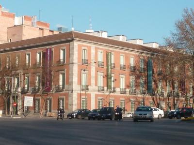 A street-level view of a large, multi-story brick building with vertical banners hanging from the facade, situated at a street corner with cars and pedestrians in the foreground.