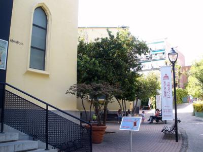 The image shows the exterior of a building with signage reading "Teatro de La Abadía" and "Absidiolo," featuring a paved courtyard, trees, benches, and people.