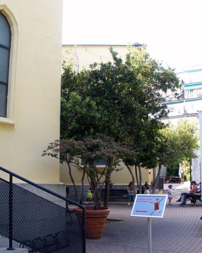 The image shows the exterior of a building with signage reading "Teatro de La Abadía" and "Absidiolo," featuring a paved courtyard, trees, benches, and people.