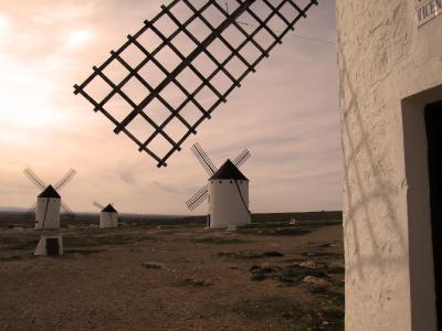 A view of several traditional white windmills with dark conical roofs and lattice sails on a dry, open plain at dusk, with the corner of a whitewashed building in the foreground right.