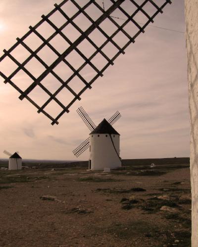 A view of several traditional white windmills with dark conical roofs and lattice sails on a dry, open plain at dusk, with the corner of a whitewashed building in the foreground right.