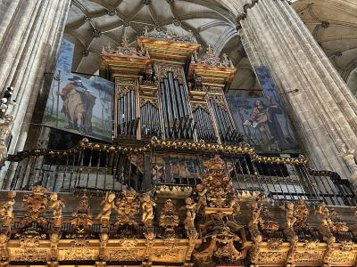 The image shows the interior of a large cathedral featuring a massive, ornate golden pipe organ and choir screen (coro) with intricate wood carvings and statues, set against high stone vaulted ceilings and columns.