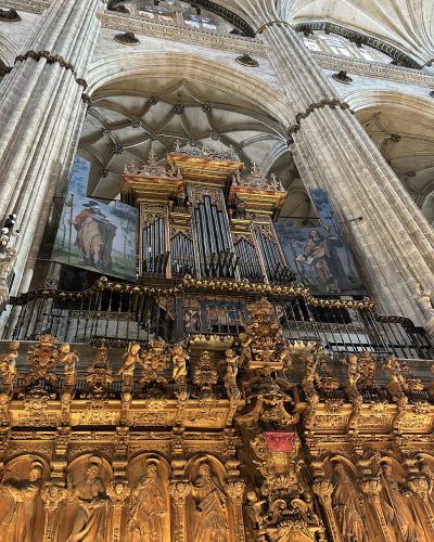 The image shows the interior of a large cathedral featuring a massive, ornate golden pipe organ and choir screen (coro) with intricate wood carvings and statues, set against high stone vaulted ceilings and columns.