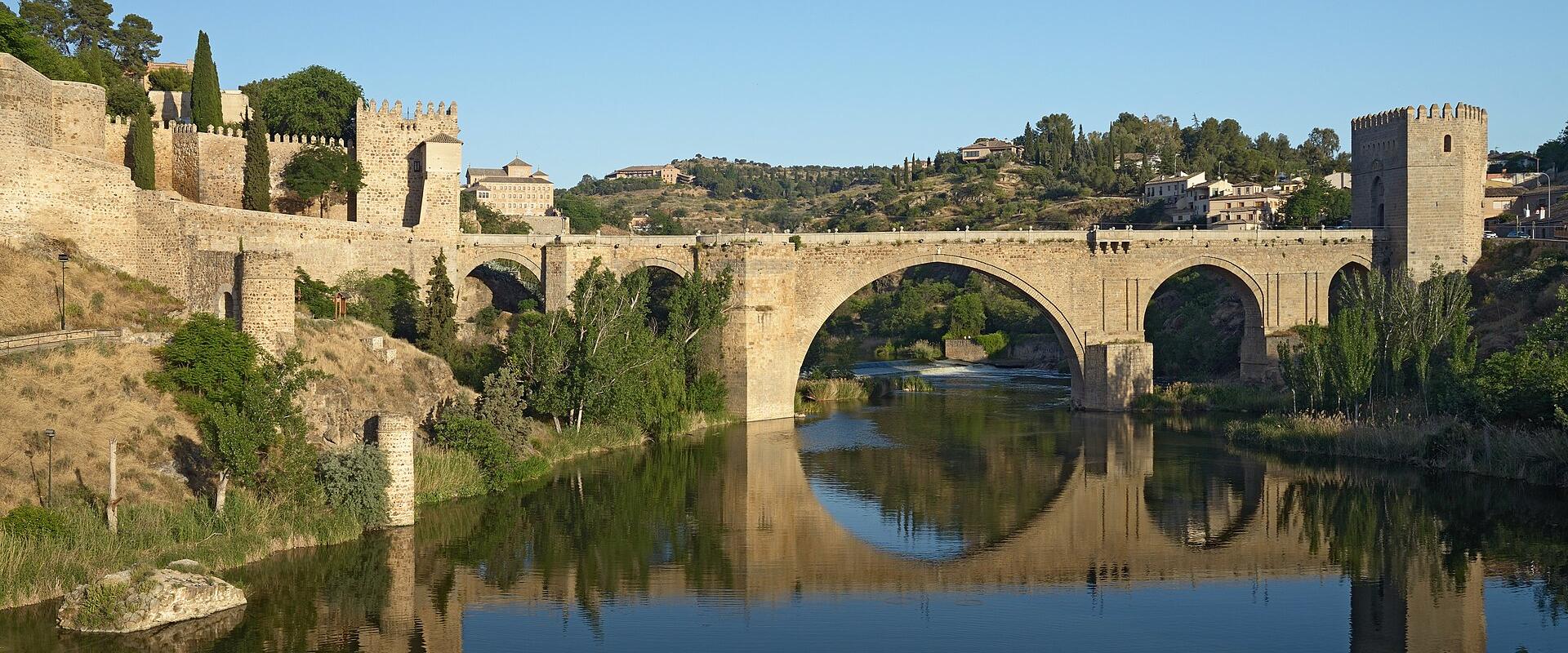 A stone arch bridge with fortified towers at either end spanning a river, with a walled city and hillside buildings in the background.