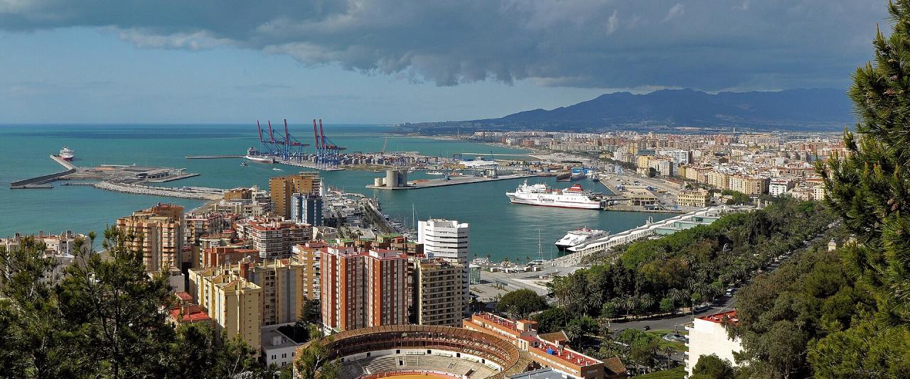 A high-angle panoramic view of a coastal city featuring a large bullring in the foreground, a busy port with container cranes and cruise ships, and a dense urban area stretching along the coastline under a cloudy sky.