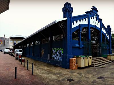 A blue, ornate metal market building with a sign reading "PESCADERIA" (fish market) on its facade, situated next to a street with parked cars and a cylindrical tower in the background.