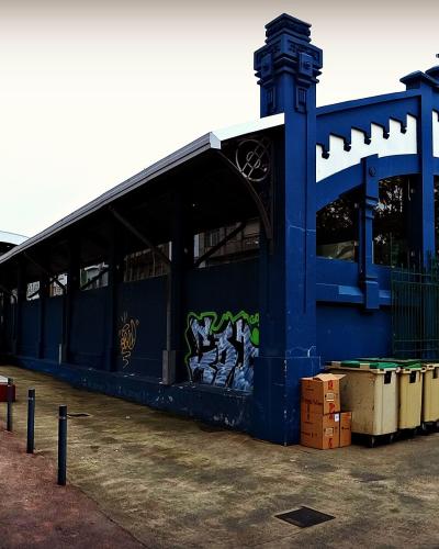 A blue, ornate metal market building with a sign reading "PESCADERIA" (fish market) on its facade, situated next to a street with parked cars and a cylindrical tower in the background.