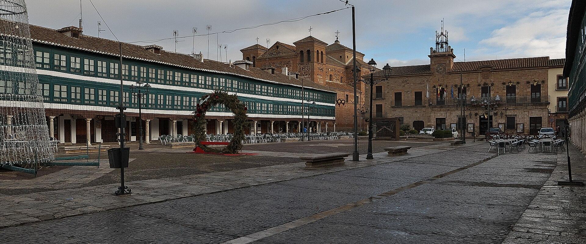 A wide, wet cobblestone plaza in Almagro, Spain, features historic buildings, a large green wreath, and outdoor café seating under an overcast sky.