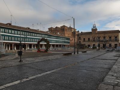 A wide, wet cobblestone plaza in Almagro, Spain, features historic buildings, a large green wreath, and outdoor café seating under an overcast sky.