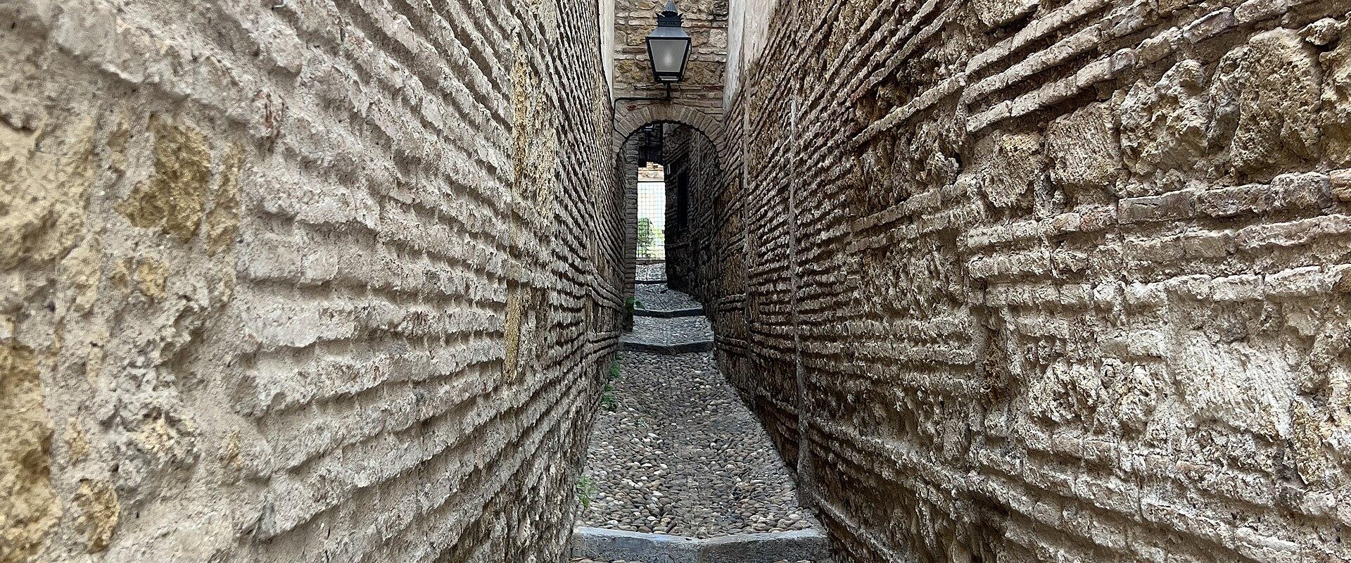 A narrow, cobblestone alleyway flanked by high walls constructed with distinctive horizontal bands of brick and stone, leading towards a series of arches.
