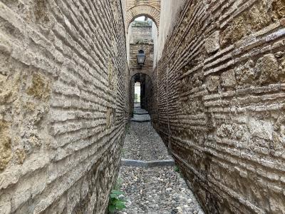 A narrow, cobblestone alleyway flanked by high walls constructed with distinctive horizontal bands of brick and stone, leading towards a series of arches.
