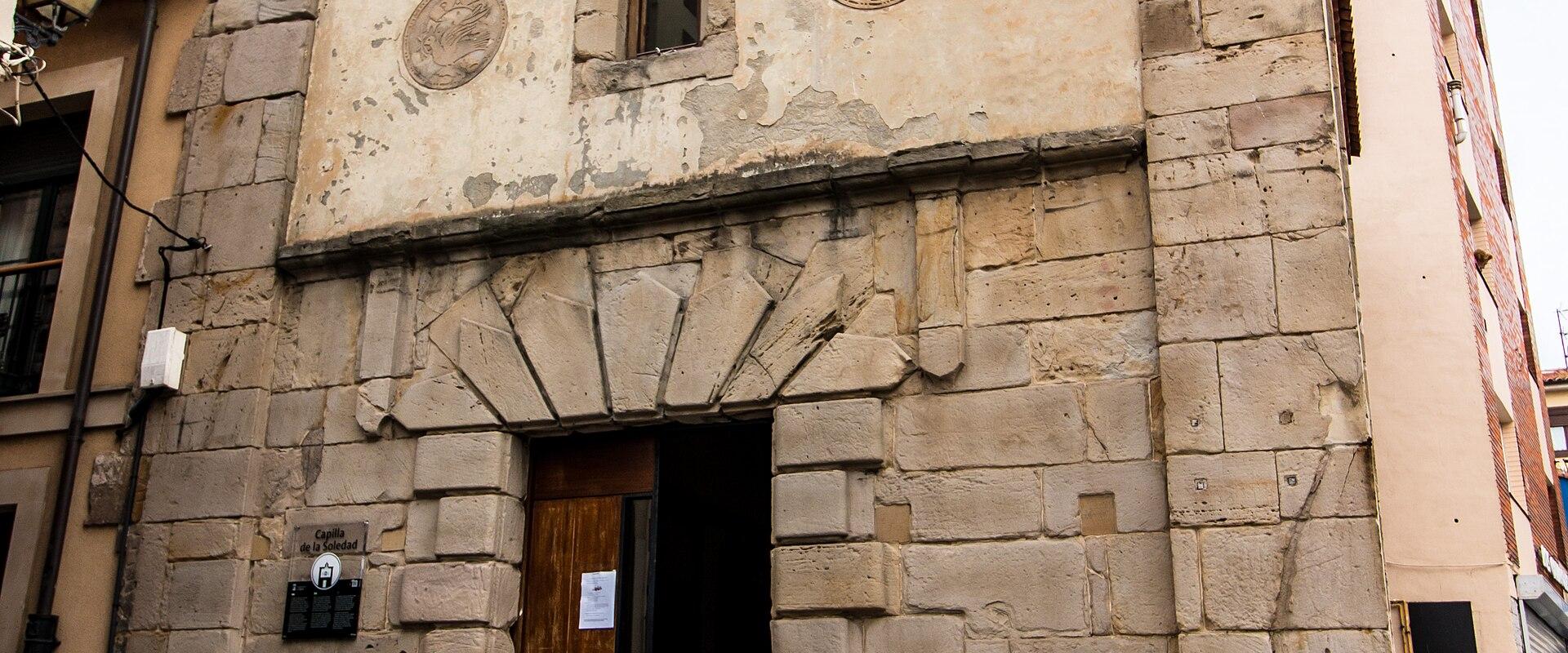 The image shows the facade of a small, stone-walled chapel with a terracotta tiled roof, a bell in a small bell tower, and a plaque on the wall to the left of the entrance that reads "Capilla de la Soledad".