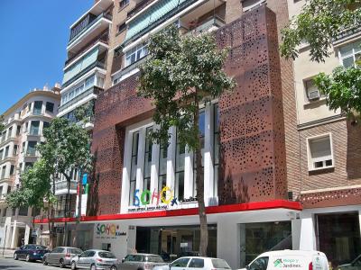 A modern multi-story building with a distinctive rust-colored perforated metal facade, featuring large glass windows and signage reading "SOHO" and "TEATRO MUSICAL BARRETA MÁLAGA" above the entrance, situated on a city street with parked cars and tre