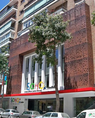 A modern multi-story building with a distinctive rust-colored perforated metal facade, featuring large glass windows and signage reading "SOHO" and "TEATRO MUSICAL BARRETA MÁLAGA" above the entrance, situated on a city street with parked cars and tre