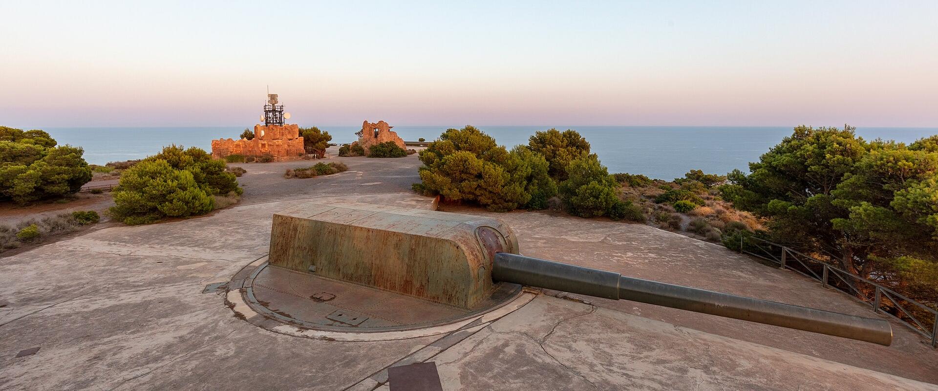A panoramic view of a coastal military fortification featuring a large naval gun in a concrete emplacement in the foreground and ruined stone walls with a lighthouse structure in the background, overlooking the sea.