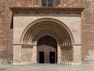 A close-up architectural view of a large, ornate stone portal with multiple concentric arches and columns, set into a weathered brick and stone wall, featuring a dark wooden double door.