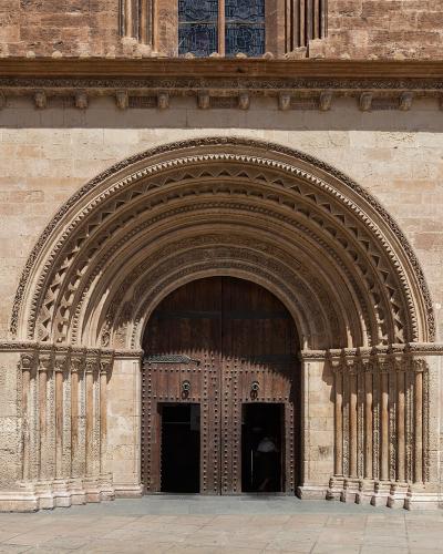 A close-up architectural view of a large, ornate stone portal with multiple concentric arches and columns, set into a weathered brick and stone wall, featuring a dark wooden double door.