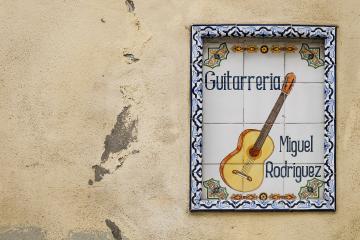 A ceramic tile sign mounted on a weathered beige wall, displaying the text "Guitarreria Miguel Rodriguez" alongside a painted image of a classical guitar.