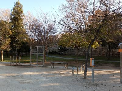 An outdoor fitness area with wooden and metal exercise equipment, a bench, and trees, situated next to a stone wall in a park-like setting.
