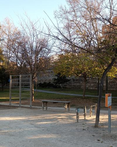 An outdoor fitness area with wooden and metal exercise equipment, a bench, and trees, situated next to a stone wall in a park-like setting.