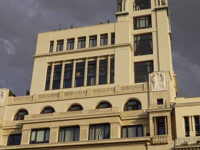 A low-angle shot of a large, cream-colored neoclassical building with a prominent central tower, columns, and relief sculptures, featuring yellow and white banners at street level advertising "PhotoESPAÑA" and "Cursos de Verano".