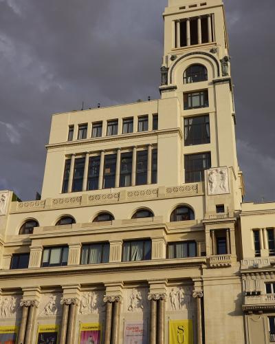 A low-angle shot of a large, cream-colored neoclassical building with a prominent central tower, columns, and relief sculptures, featuring yellow and white banners at street level advertising "PhotoESPAÑA" and "Cursos de Verano".