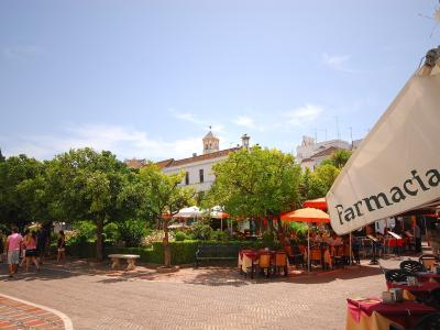 A sunlit public square with orange trees, paved walkways, outdoor café seating under umbrellas, and a white building with a bell tower in the background; a large white umbrella in the foreground displays the word “Farmacia” with a green cross symbol.