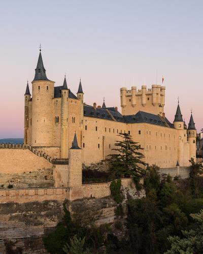 A large medieval stone castle with distinctive conical slate-roofed towers and a fortified keep, situated on a rocky promontory overlooking a town with a prominent cathedral spire in the background.