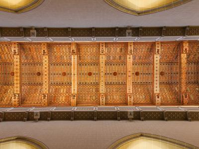 An interior view looking up at an ornate, coffered wooden ceiling (techumbre) with geometric and floral patterns, supported by decorative beams, inside a large cathedral-like space.