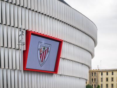The image shows the exterior of a modern stadium with a distinctive white, curved facade and a large digital screen displaying the crest of Athletic Club Bilbao.