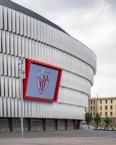 The image shows the exterior of a modern stadium with a distinctive white, curved facade and a large digital screen displaying the crest of Athletic Club Bilbao.