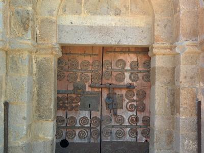 A close-up of a Romanesque stone doorway with a wooden double door adorned with spiral ironwork, set within a rounded archway.