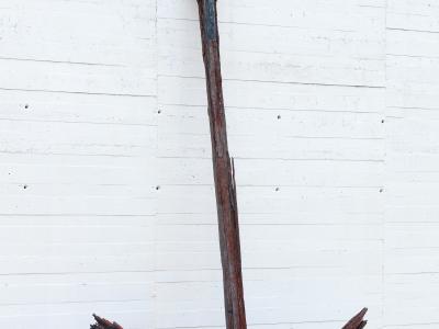 A large, heavily rusted and weathered wooden anchor standing upright against a white concrete wall with horizontal formwork lines, on a cobblestone surface.