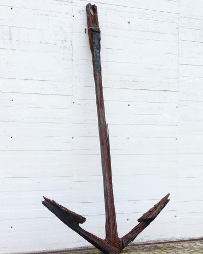 A large, heavily rusted and weathered wooden anchor standing upright against a white concrete wall with horizontal formwork lines, on a cobblestone surface.