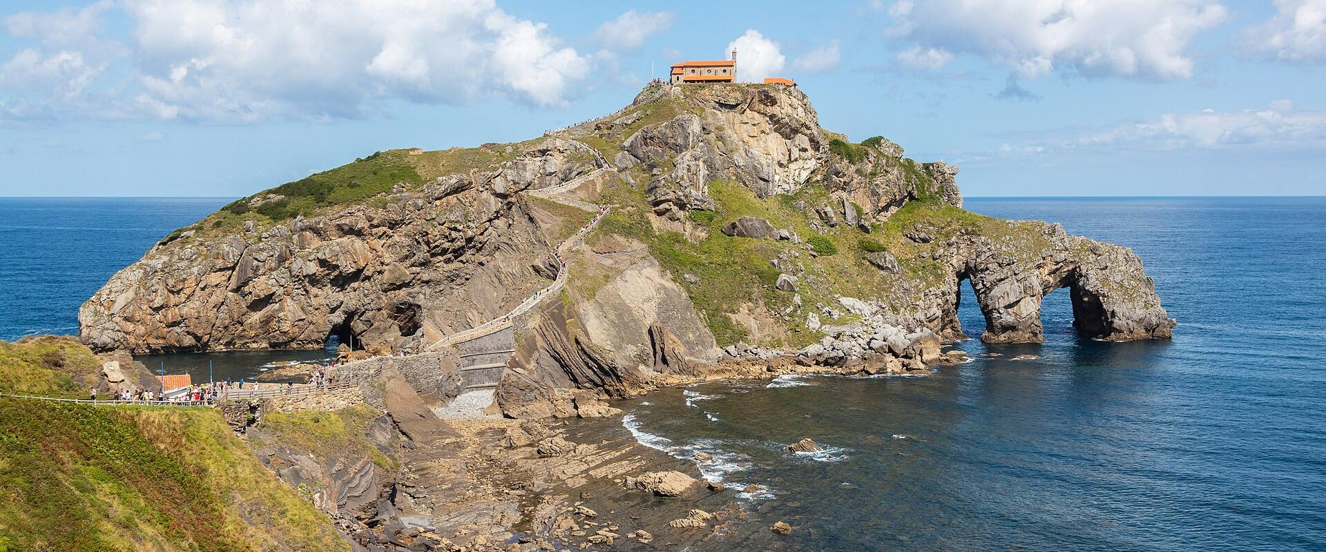 A rocky coastal islet connected to the mainland by a winding stone staircase, featuring a small hermitage at its peak and a natural sea arch at its base, with tourists visible on the path.