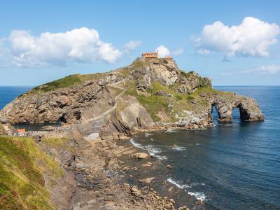A rocky coastal islet connected to the mainland by a winding stone staircase, featuring a small hermitage at its peak and a natural sea arch at its base, with tourists visible on the path.