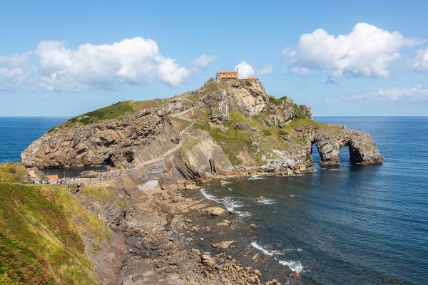 A rocky coastal islet connected to the mainland by a winding stone staircase, featuring a small hermitage at its peak and a natural sea arch at its base, with tourists visible on the path.