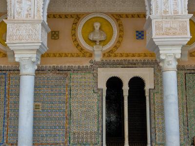 The image shows an ornate interior courtyard featuring white horseshoe arches with intricate stucco carvings, walls covered in colorful geometric tiles (azulejos), and a bust of a classical figure set within a yellow circular niche.