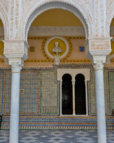 The image shows an ornate interior courtyard featuring white horseshoe arches with intricate stucco carvings, walls covered in colorful geometric tiles (azulejos), and a bust of a classical figure set within a yellow circular niche.