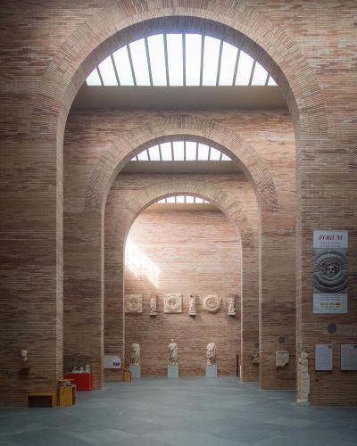 The image shows the interior of a modern museum with distinctive red brick arches, skylights, and Roman sculptures on display.
