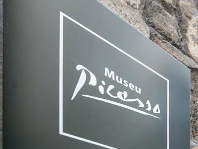 A close-up of a dark grey directional sign mounted on a stone wall, featuring the white text "Museu Picasso" in a stylized font and "Ajuntament de Barcelona" at the bottom.