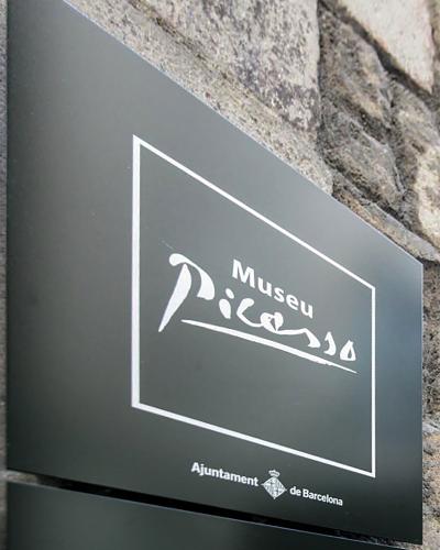 A close-up of a dark grey directional sign mounted on a stone wall, featuring the white text "Museu Picasso" in a stylized font and "Ajuntament de Barcelona" at the bottom.