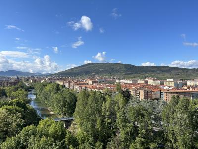 A scenic overlook of a city with dense green trees in the foreground, a river flowing through the middle ground, and a large hill rising behind a cluster of buildings with terracotta roofs under a blue sky.