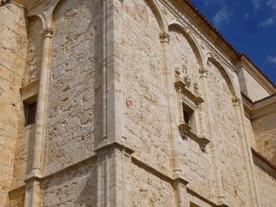 A low-angle shot of the corner of a light-colored stone church facade featuring Gothic-style blind arches, decorative columns, and a small window with a sculpted frame under a blue sky.