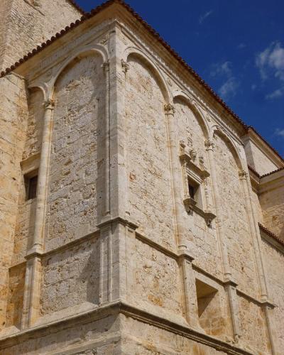 A low-angle shot of the corner of a light-colored stone church facade featuring Gothic-style blind arches, decorative columns, and a small window with a sculpted frame under a blue sky.