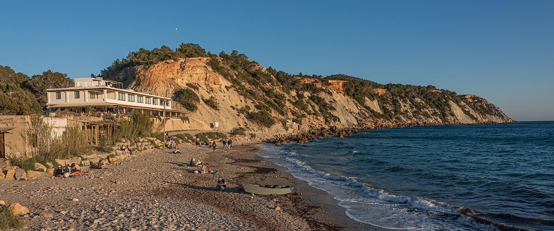 A coastal scene showing a pebble beach, a white multi-story building built into a cliffside, and the sea under a clear blue sky.